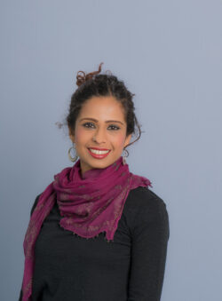 A headshot of Yamini Narayanan, a woman with dark hair styled in a bun, wearing a black top, a vibrant purple scarf with small patterns, and large silver hoop earrings, smiling against a light blue background.