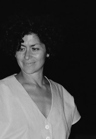 An artistic, black-and-white portrait of a woman with dark, voluminous curly hair looking slightly downward and to her left with a faint, contemplative smile. She is wearing a light-colored, V-neck garment with visible buttons. The background is completely dark, creating a high-contrast, moody aesthetic that focuses entirely on her expression and the texture of her hair.