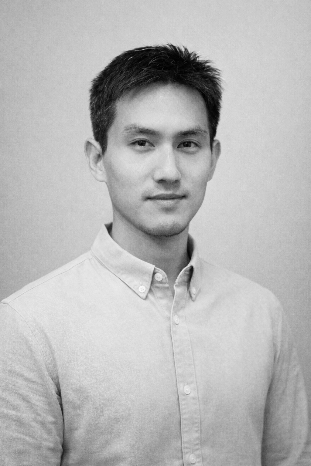 A black and white, waist-up portrait of a young man with short, dark hair and a light goatee. He is looking directly at the camera with a neutral, calm expression. He is wearing a light-colored, button-down collared shirt. The background is a solid, neutral tone with a soft, textured appearance. The lighting is soft and even, highlighting his features.