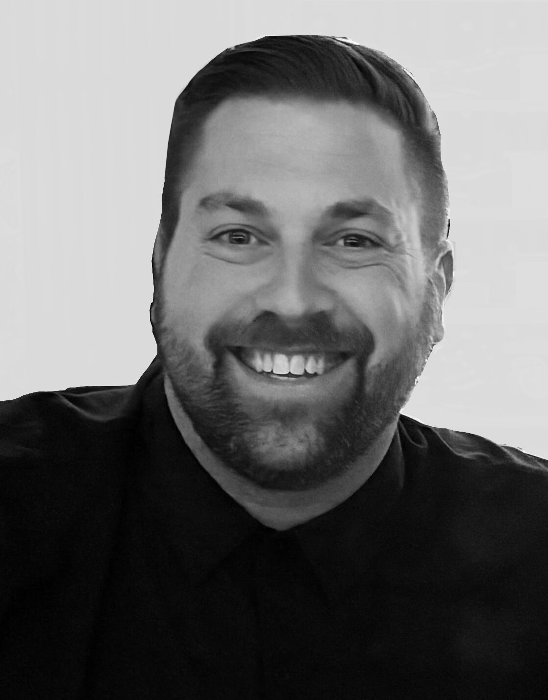A black and white, close-up headshot of a smiling man with short, styled dark hair and a well-groomed beard and mustache. He is wearing a dark, button-down shirt and is positioned against a plain, light-gray background. The man is looking directly at the camera with a warm and friendly expression.
