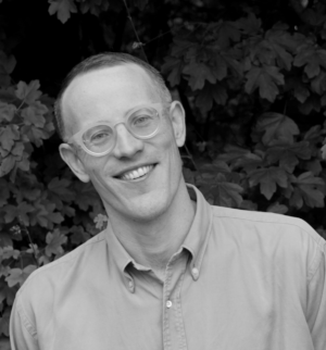 A black and white, square-format headshot of a smiling man with short hair and light-colored, round-framed glasses. He is wearing a button-down shirt and standing in front of a background of leafy foliage.