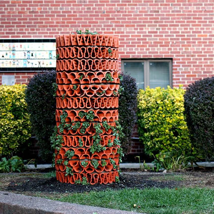 Here is the alt text for your images, categorized by file name: BIRKELAND-HEADSHOT.png Standard: A black and white, close-up headshot of a woman with dark hair pulled back, smiling warmly at the camera. She wears long, thin drop earrings and a dark V-neck top. Concise: Black and white portrait of a smiling woman with dark hair. jb-work-3.jpg Standard: A large, rounded hut made of dried grass and hay stands in a grassy field. A jagged opening in the front reveals a vibrant red and purple patterned interior. To the right, a person with a backpack stands in profile, looking toward the structure. Concise: A hay-covered hut with a colorful interior opening in a wooded field. jb-work-4.jpg Standard: An architectural installation in a courtyard with large stone pillars and arched ceilings. A geometric, tree-like pattern made of small white circular elements is mounted on the pillars, branching upward and outward across the stone surfaces. Concise: Tree-like geometric installation on stone pillars in a vaulted courtyard. jb-work-2.jpg Standard: A top-down view of a modular planter system made of interlocking terracotta-colored hexagonal and triangular tiles. Some tiles contain small green succulents or clusters of vibrant pink flowers, while others remain empty, showing white internal structures. Concise: Top-down view of a terracotta-colored modular planter with pink flowers and succulents. jb-work-.jpg Standard: A tall, cylindrical planter column made of stacked, wavy terracotta tiles sits in a garden. Small green plants grow from the openings of the tiles. In the background are green and dark purple bushes in front of a red brick building.