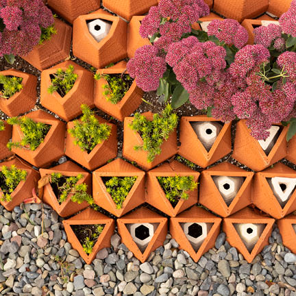 A top-down view of a modular planter system made of interlocking terracotta-colored hexagonal tiles. Some tiles contain small green succulents or clusters of vibrant pink flowers, while others remain empty, showing white internal structures.