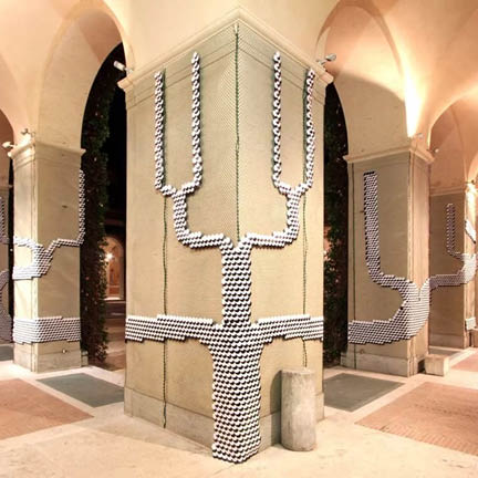 An architectural installation in a courtyard featuring large stone pillars under arched ceilings. A geometric, tree-like pattern made of small white circular elements is mounted onto the surface of the pillars, branching upward and outward.