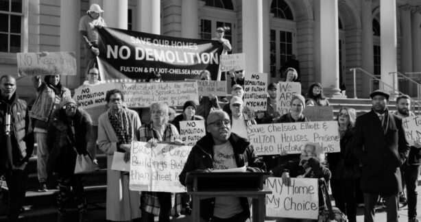 A black-and-white photo of a protest on building steps against the demolition of public housing in Chelsea. A woman speaks at a lectern, backed by a diverse crowd holding signs and a central banner reading: 