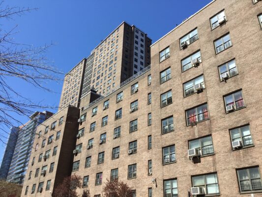 A low-angle shot of several tan brick public housing apartment buildings against a clear blue sky. The buildings vary in height, with numerous windows and window-mounted air conditioning units visible across the facades.