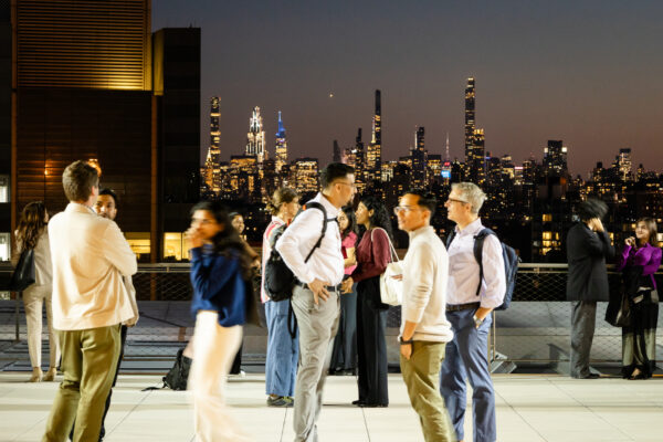 A medium shot captures a group of people socializing during an evening event on an outdoor rooftop terrace. The foreground and middle ground show various individuals in semi-formal and business-casual attire—some standing in small groups, others walking—rendered with a slight motion blur that suggests a lively atmosphere. In the background, the illuminated New York City skyline stretches across the horizon under a dusky, dark blue sky, featuring prominent skyscrapers and city lights. The terrace is paved with light-colored square tiles, and a safety cable railing runs along the edge, separating the guests from the urban backdrop.