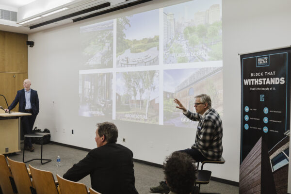 A man in a plaid shirt sits on a stool, gesturing with his hand as he speaks during a presentation.