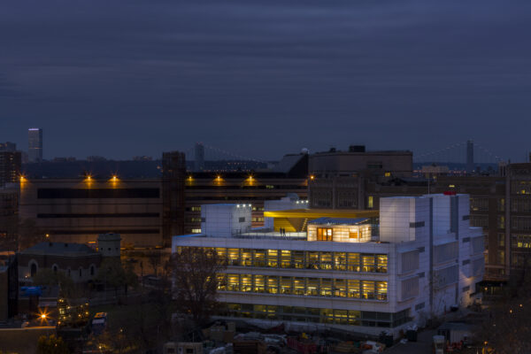 A wide landscape shot at twilight features a modern, brightly lit white building in the center foreground, surrounded by an urban environment. The building's interior glow highlights its grid-like window pattern, while a distinct, glowing yellow structure and a smaller rectangular 