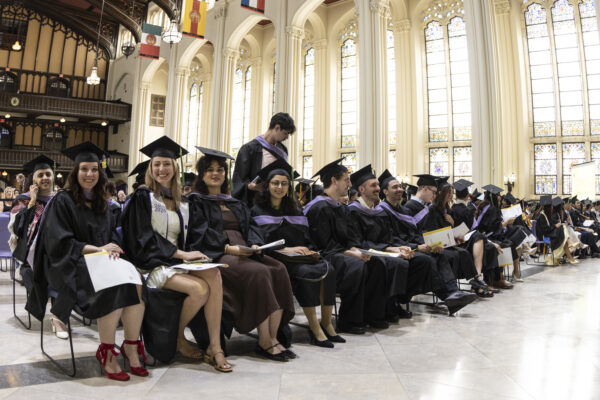 A row of seated graduates in black caps and gowns smiling during a ceremony inside a grand hall with stained-glass windows.