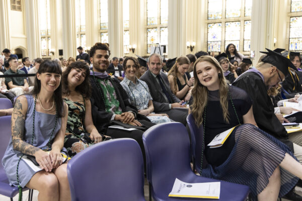A group of smiling guests and a graduate seated in purple chairs among the audience at a graduation event.