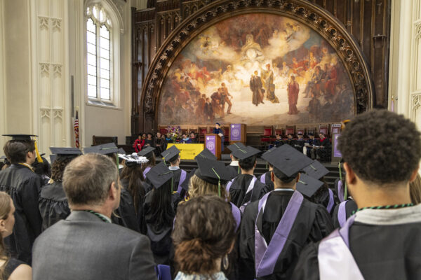 The view from behind the seated graduates looking toward a speaker at the podium on a stage featuring a large mural.