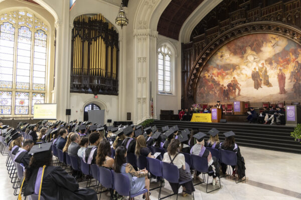 A wide angle from behind the audience of graduates watching a speaker on stage in a hall with a massive mural and pipe organ.