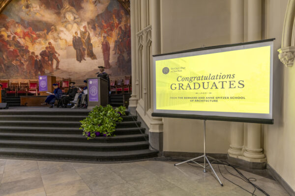 A projector screen displaying a congratulatory message for graduates of the Spitzer School of Architecture, with the stage in the background.