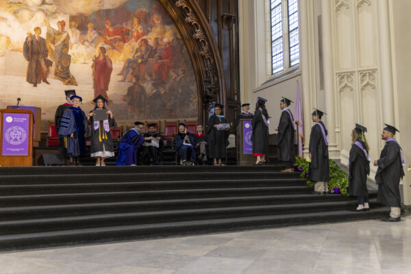 Graduates walking across a stage to receive their diplomas from faculty members in front of a large mural.