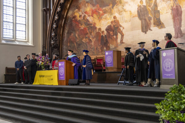 A speaker addressing the audience from a podium on stage alongside faculty members during the graduation ceremony.