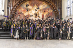 A large group of joyous graduates throwing their caps into the air while posing on steps in front of a grand mural.