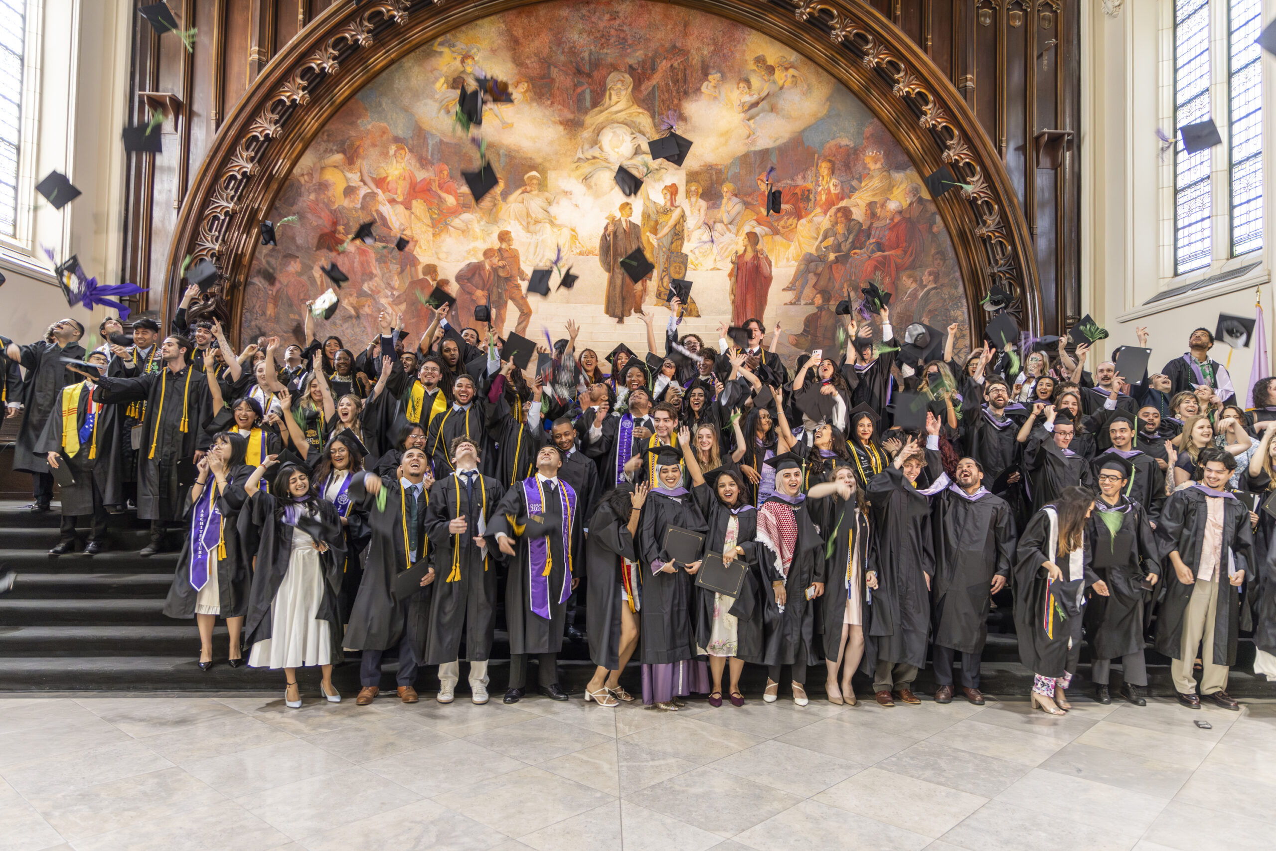 A large group of joyous graduates throwing their caps into the air while posing on steps in front of a grand mural.