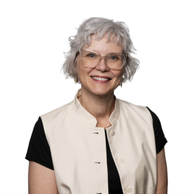A professional headshot of a smiling woman with short, curly silver hair and clear-rimmed glasses, wearing a black shirt under a cream-colored button-down vest against a white background.