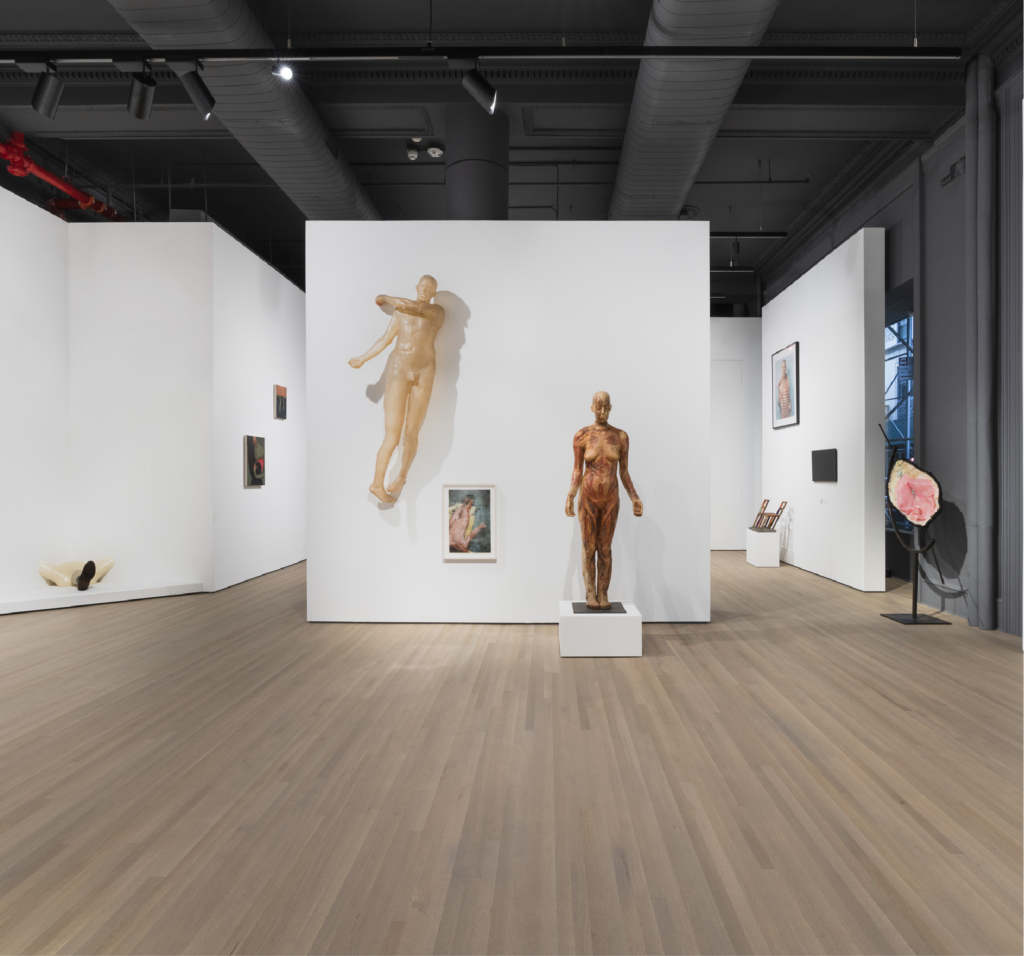 Wide interior photograph of an expansive, contemporary art gallery space with light-toned wood plank flooring and an open, charcoal-painted industrial ceiling with exposed ducts and track lighting. Centered in the foreground is a white partition wall displaying a life-sized sculpture of a standing anatomical female figure on a low white plinth, alongside a wall-mounted male figure positioned diagonally as if suspended or floating, and a small framed artwork below it. Adjacent gallery walls lead into adjoining spaces, displaying additional framed art pieces, small pedestals, and a freestanding sculpture.