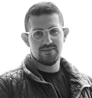 Black-and-white portrait of a man with short dark hair, a trimmed beard, and mustache, looking directly forward with a gentle smile. He is wearing round, clear or light-colored eyeglasses, a dark crew-neck shirt, and an unbuttoned quilted patterned jacket against a bright, softly lit background.