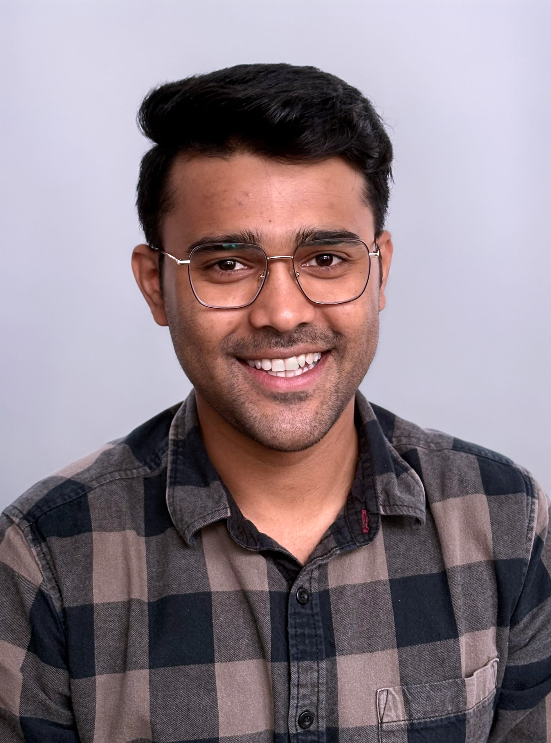A headshot of a smiling young man with dark hair, wireframe glasses, and light facial hair, wearing a brown and black plaid button-down shirt against a solid light gray background.