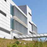 An exterior view of The Bernard and Anne Spitzer School of Architecture at the City College of New York under a clear blue sky. The modern white panel building features rows of vertical glass windows shaded by external louvered metal screens, with a grassy slope and wildflowers in the foreground. An enclosed glass walkway extends outward, displaying the school's name along its structural frame.