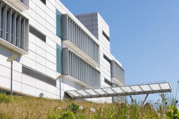 An exterior view of The Bernard and Anne Spitzer School of Architecture at the City College of New York under a clear blue sky. The modern white panel building features rows of vertical glass windows shaded by external louvered metal screens, with a grassy slope and wildflowers in the foreground. An enclosed glass walkway extends outward, displaying the school's name along its structural frame.