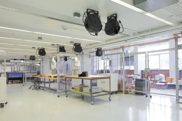 An interior view of a bright, modern laboratory space featuring robotic arms mounted on workbenches. Retractable cord reels hang from the white ceiling, and the workspace is organized with modular metal framing, glass partition walls, and equipment storage areas.